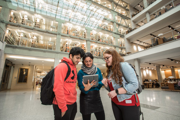 Three students standing together in a modern university atrium, looking at a tablet and collaborating on a project.